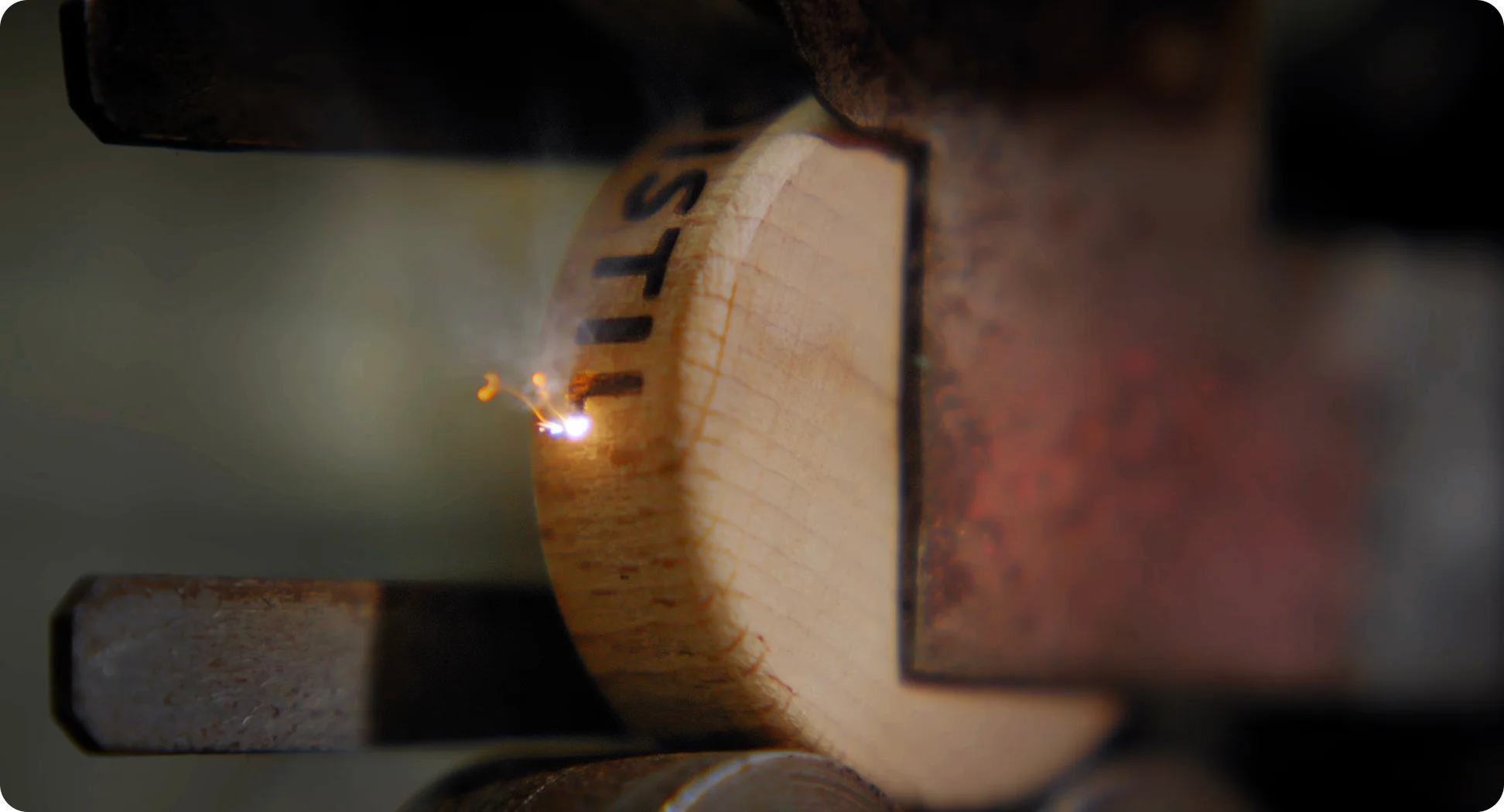 Close-up of laser engraving the word 'Talis' on a piece of light-colored wood held by metal clamps.