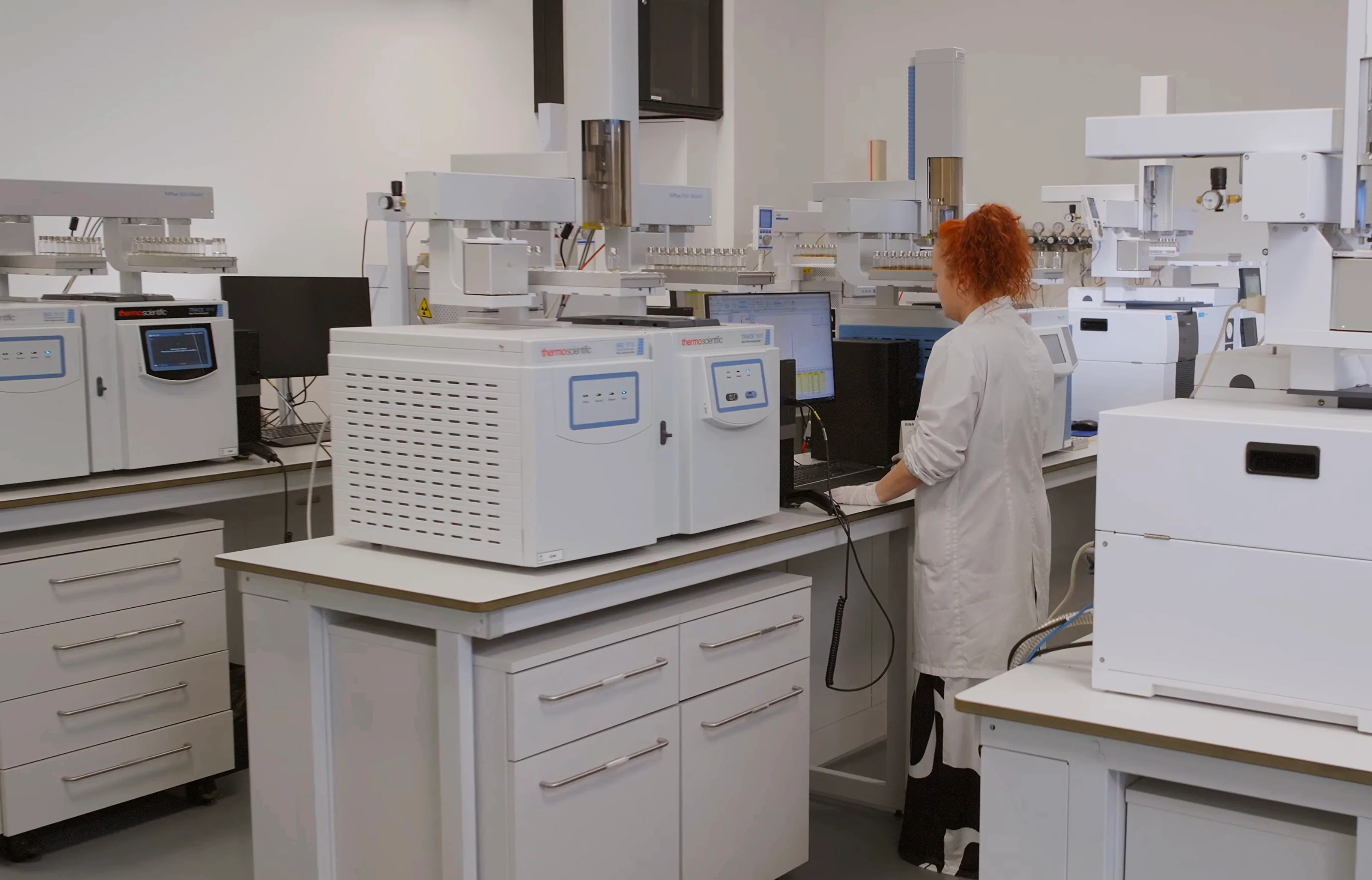 A laboratory technician in a white lab coat working on a computer surrounded by white scientific instruments and analyzers on countertops in a clean lab environment.