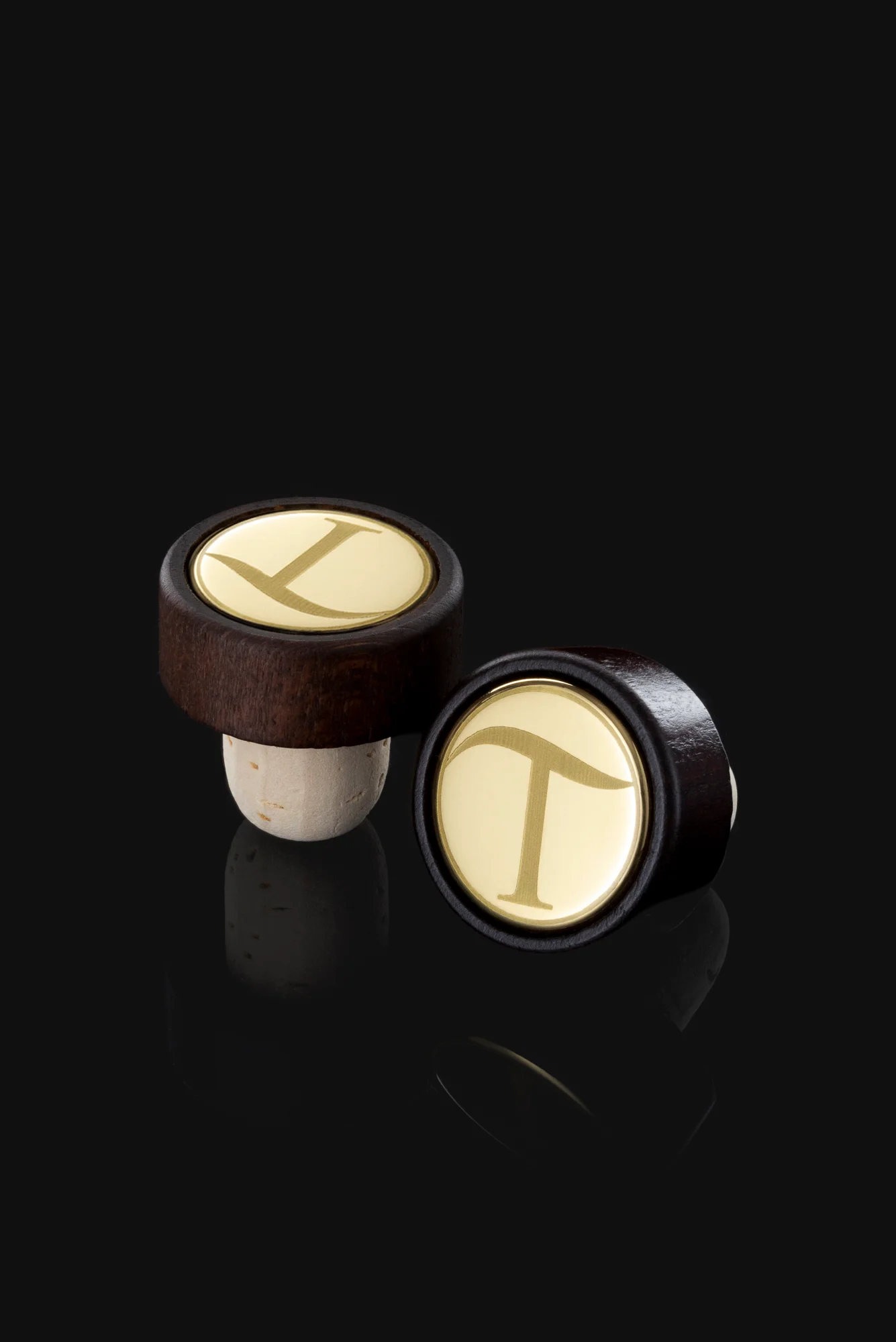 Two wine bottle stoppers with dark wood tops and golden circular caps engraved with the letter 'T' on a black background.