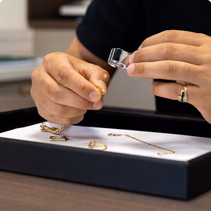 Une personne inspecte une bague en or avec une loupe sur un plateau avec plusieurs bijoux en or.
