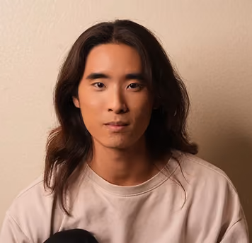 Portrait of a young person with long dark hair wearing a beige shirt against a plain wall.