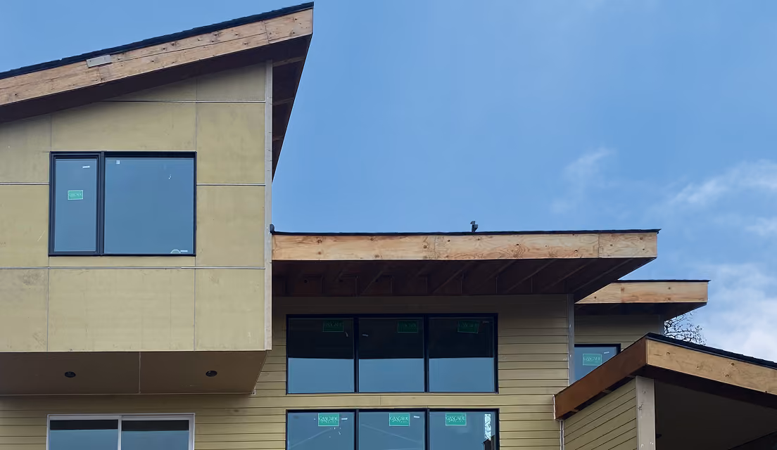Part of a modern house under construction with large windows and an angled roof under a clear blue sky.
