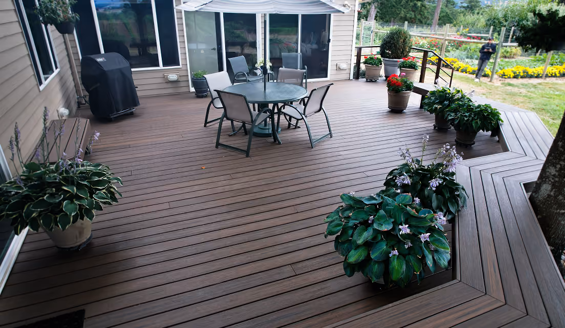 Spacious brown wooden deck with round table, chairs, umbrella, potted plants, and a grill near a house.