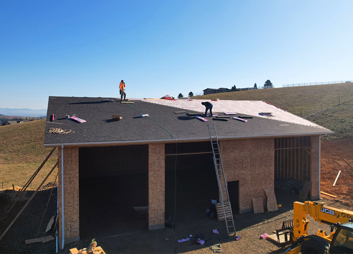 Two workers installing roofing shingles on a large wooden structure under clear blue sky.
