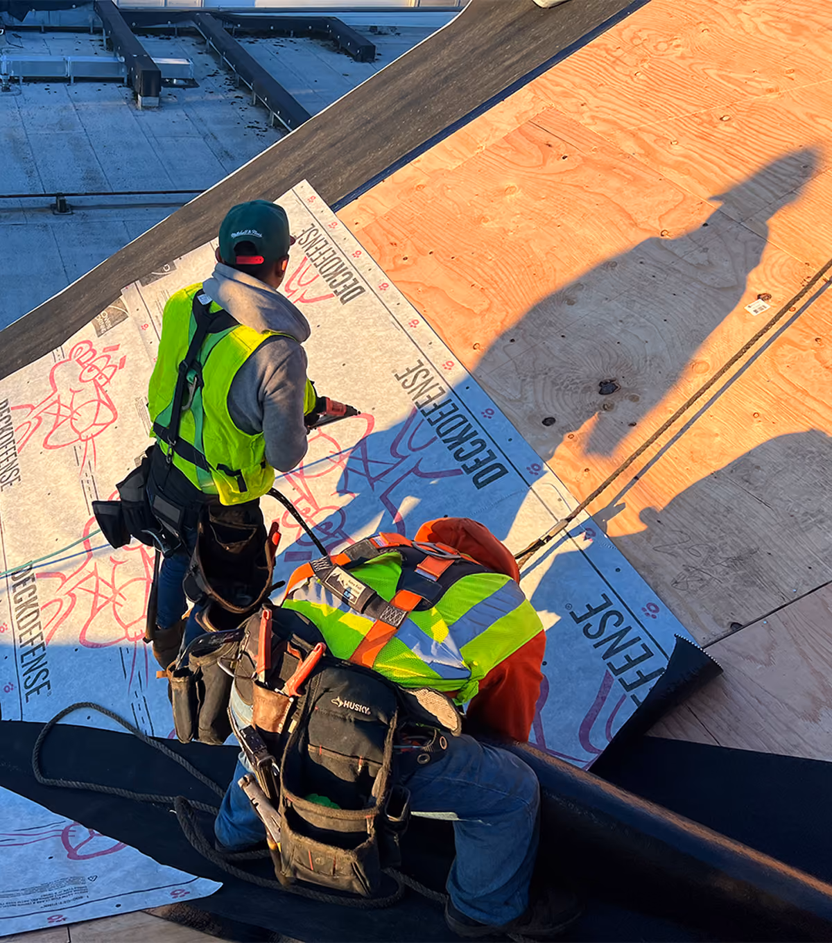 Two construction workers in safety vests installing roofing materials on a wooden roof deck.