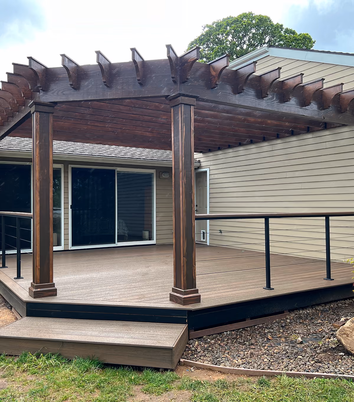 Wooden deck with dark wood pergola, black railing, and sliding glass doors on a beige house.