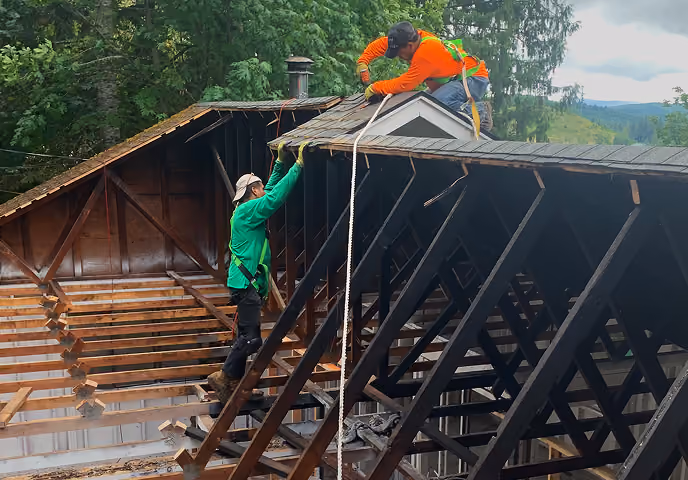 Two construction workers securing a rope on a wooden roof frame outdoors surrounded by trees.