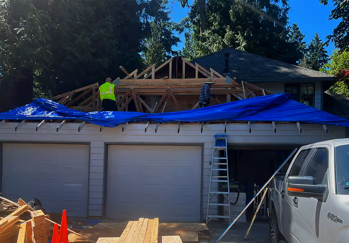 Two construction workers on roof framing covered with blue tarp above a garage and parked truck.