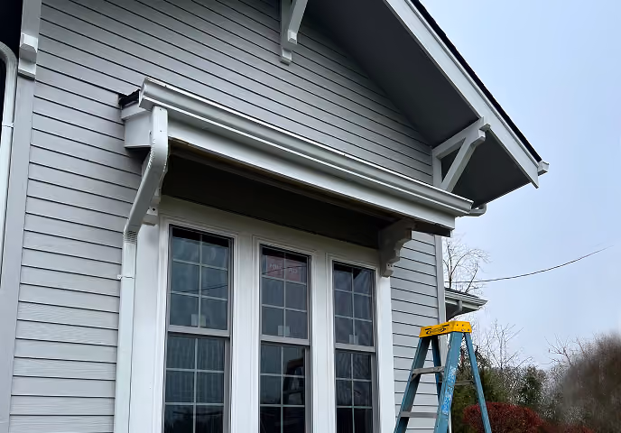 Exterior gray house wall with white-trimmed triple window and blue step ladder outdoors.