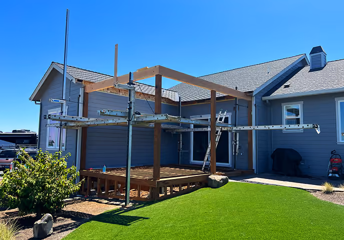 Wooden deck under construction with metal scaffolding outside a gray house under blue sky.