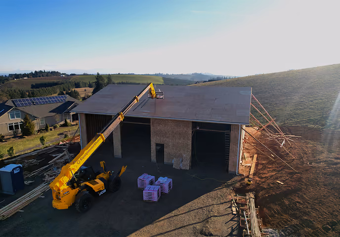 Yellow crane lifts a worker atop a partially built wooden structure on a sunny rural construction site.