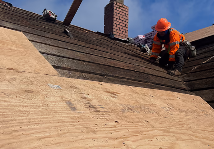 Construction worker in orange safety gear repairing a roof with wooden sheathing and old shingles.