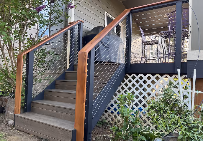 Outdoor stairs with wooden handrails and cable railing leading to a deck with metal chairs and table.