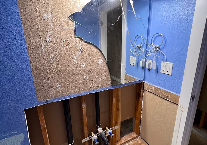 Partially demolished bathroom wall revealing plumbing pipes and a broken wall mirror above it.