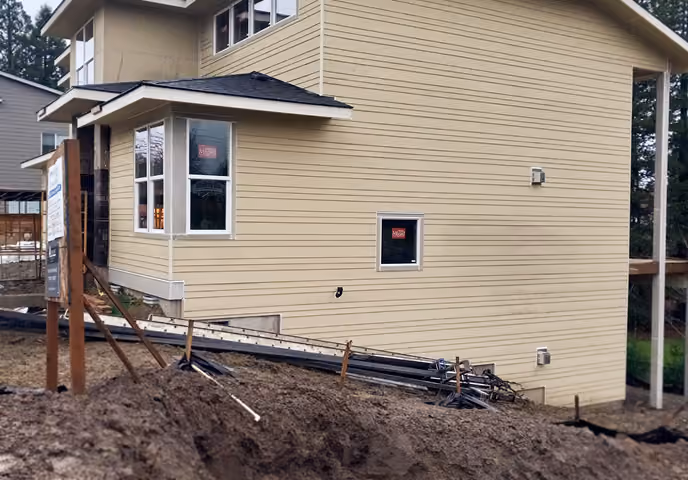 New two-story beige house with horizontal siding, construction materials, and dirt mound in front.