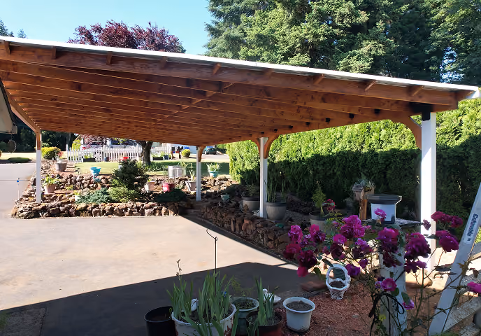 Wooden carport with stone-bordered garden beds and various potted plants under bright daylight.