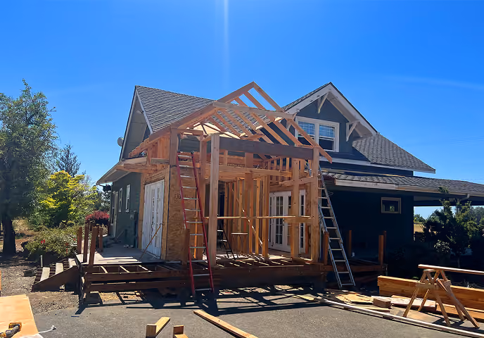 Wooden frame structure of a house extension under construction beside an existing home on a sunny day.