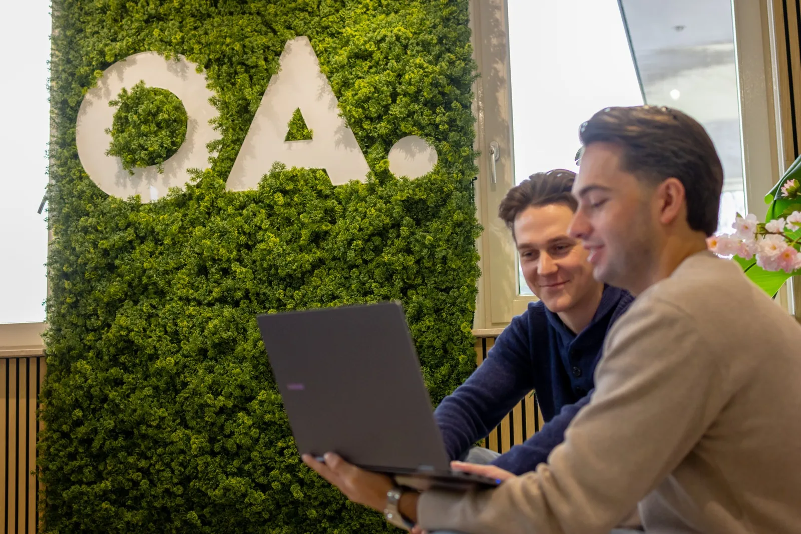 Twee collega's kijken samen naar een laptop bij de groene wand met het logo van Online Actief