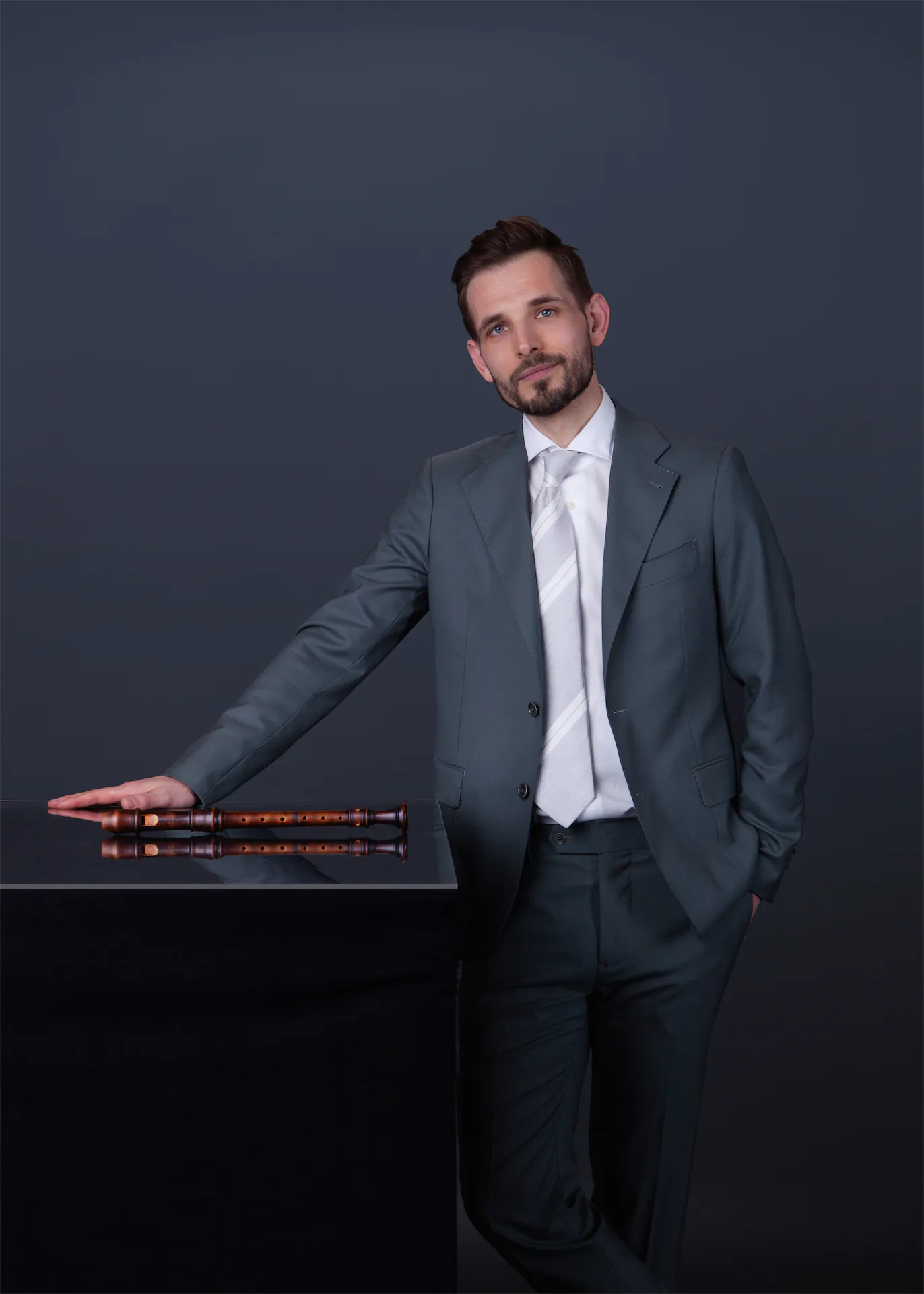 Man in suit sitting on stool playing a wooden recorder against a dark background.