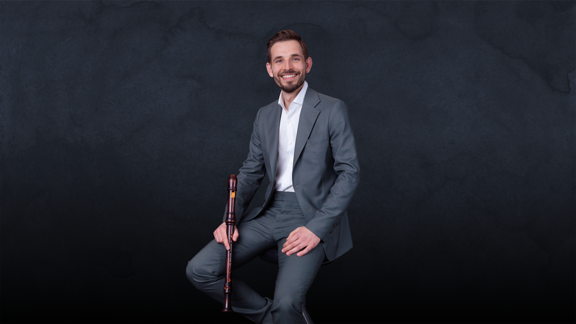 Smiling man in a grey suit sitting against dark background holding a wooden recorder instrument.