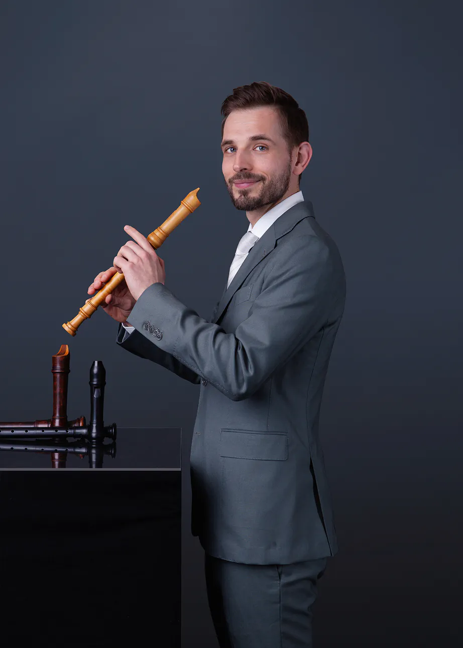 Man in gray suit holding a wooden recorder, standing next to a black surface with two other recorders.