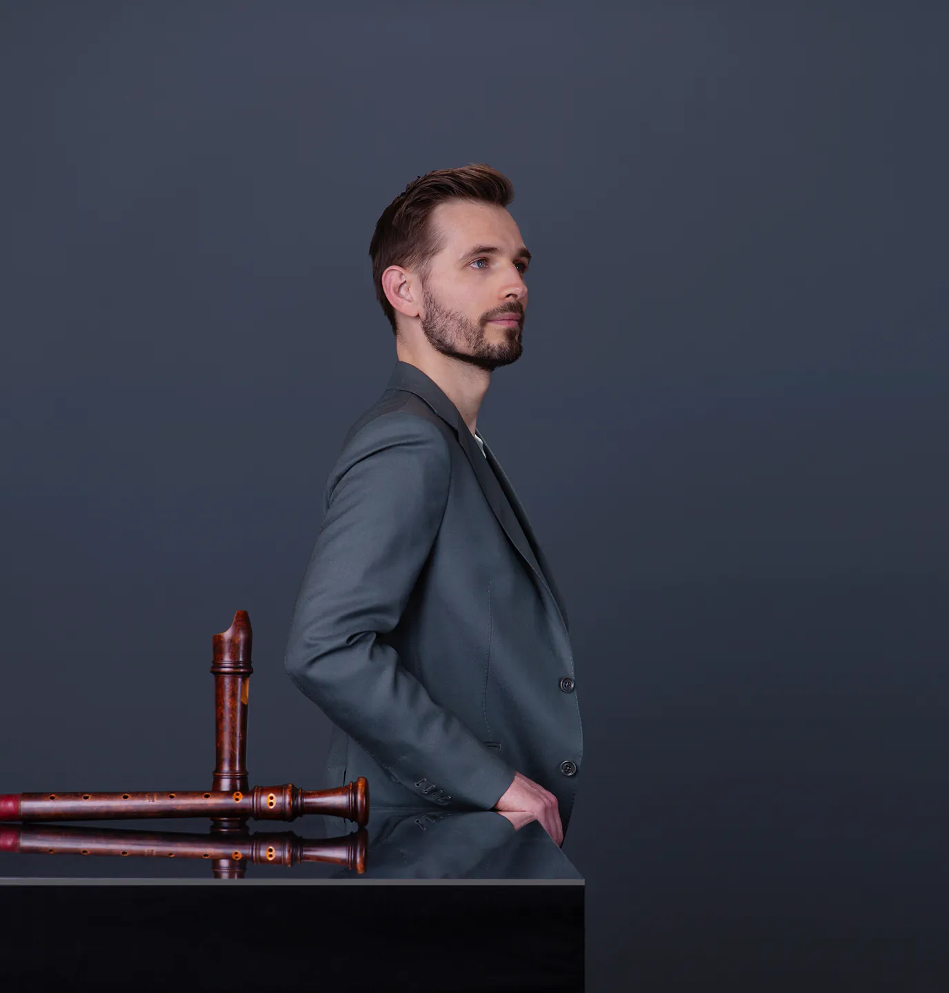Man in gray jacket standing beside a reflective black surface with two wooden recorders on it.