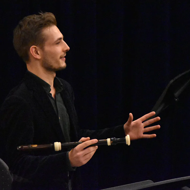 Man in black jacket holding a wooden instrument while gesturing with his other hand on stage.