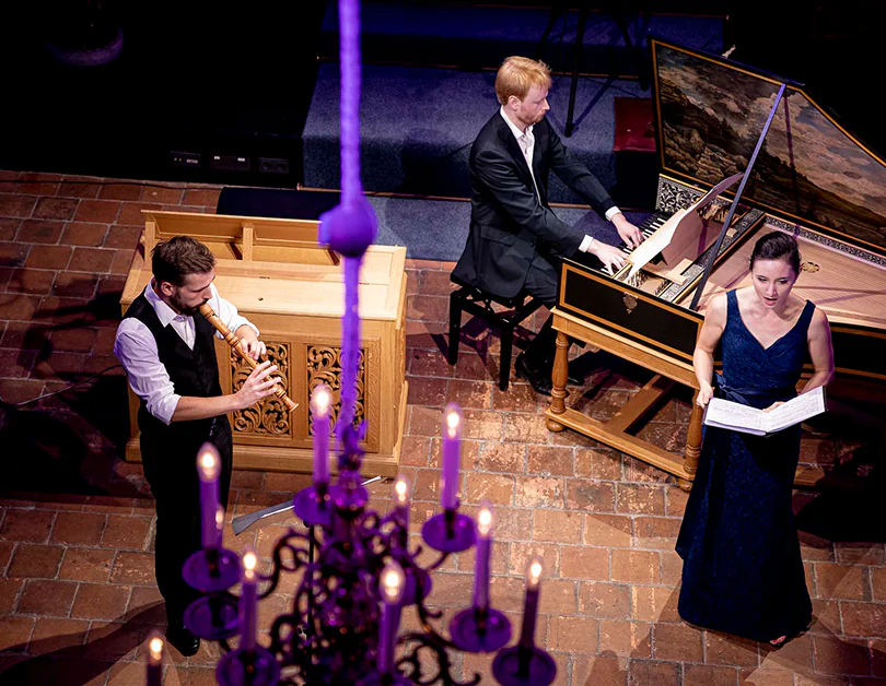 Three musicians performing indoors: man on recorder, man on harpsichord, woman singing with sheet music.