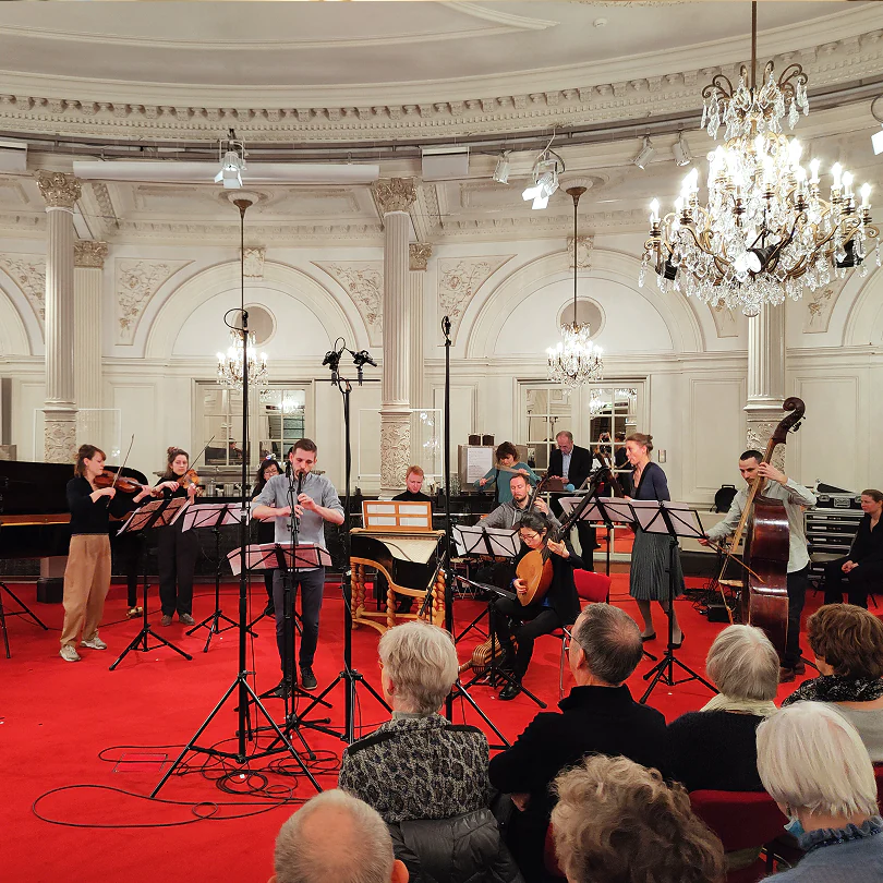 Orchestra performing in an elegant hall with chandeliers and audience seated on red carpet.