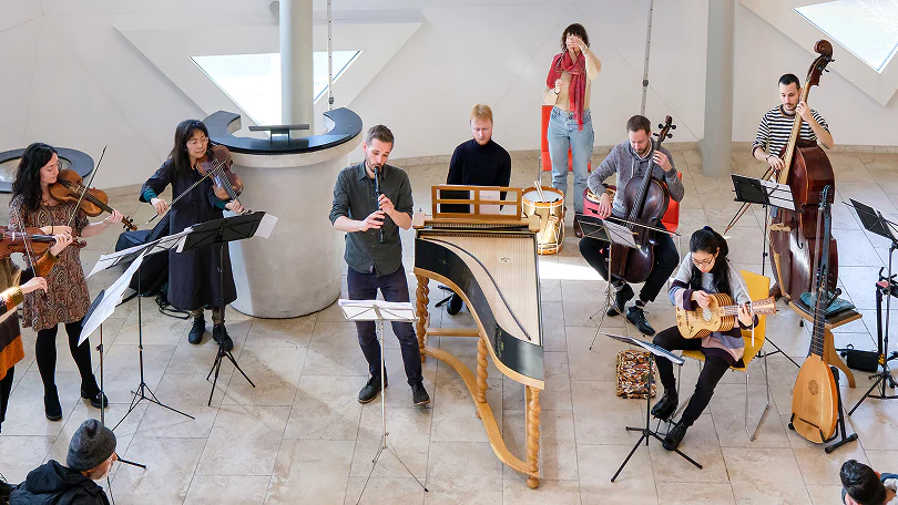 Group of musicians playing various string and wind instruments in a bright room with tile floor.