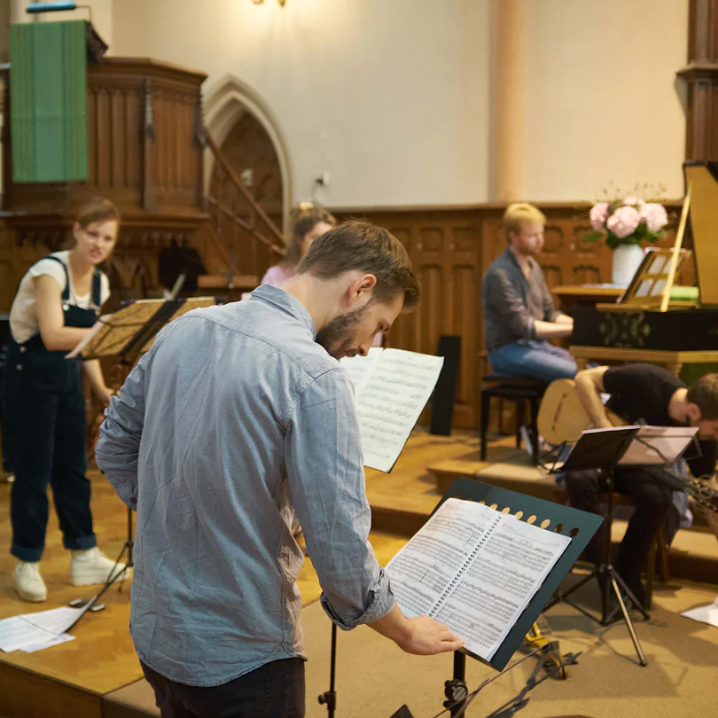 Group of musicians reading sheet music practicing in a room with wooden walls and a piano.