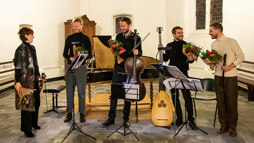 Five musicians holding flowers and standing in front of a harpsichord in a performance space.