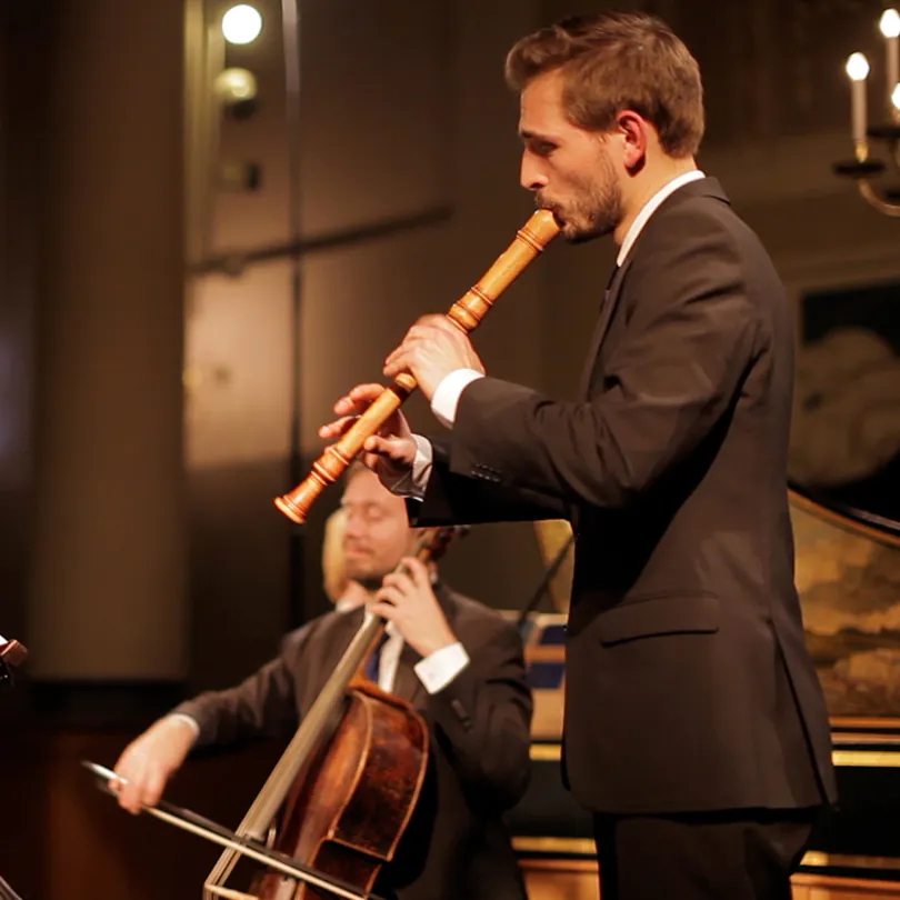 Man in suit playing wooden recorder with another musician playing cello in background.