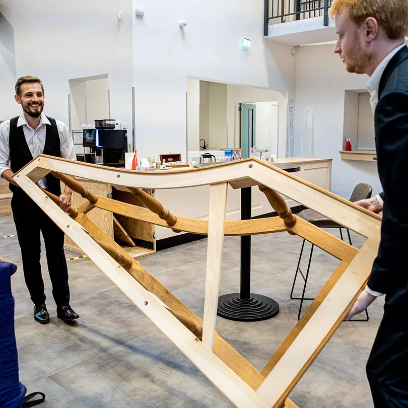 Two men in suits lifting a large wooden frame inside a modern, bright space.