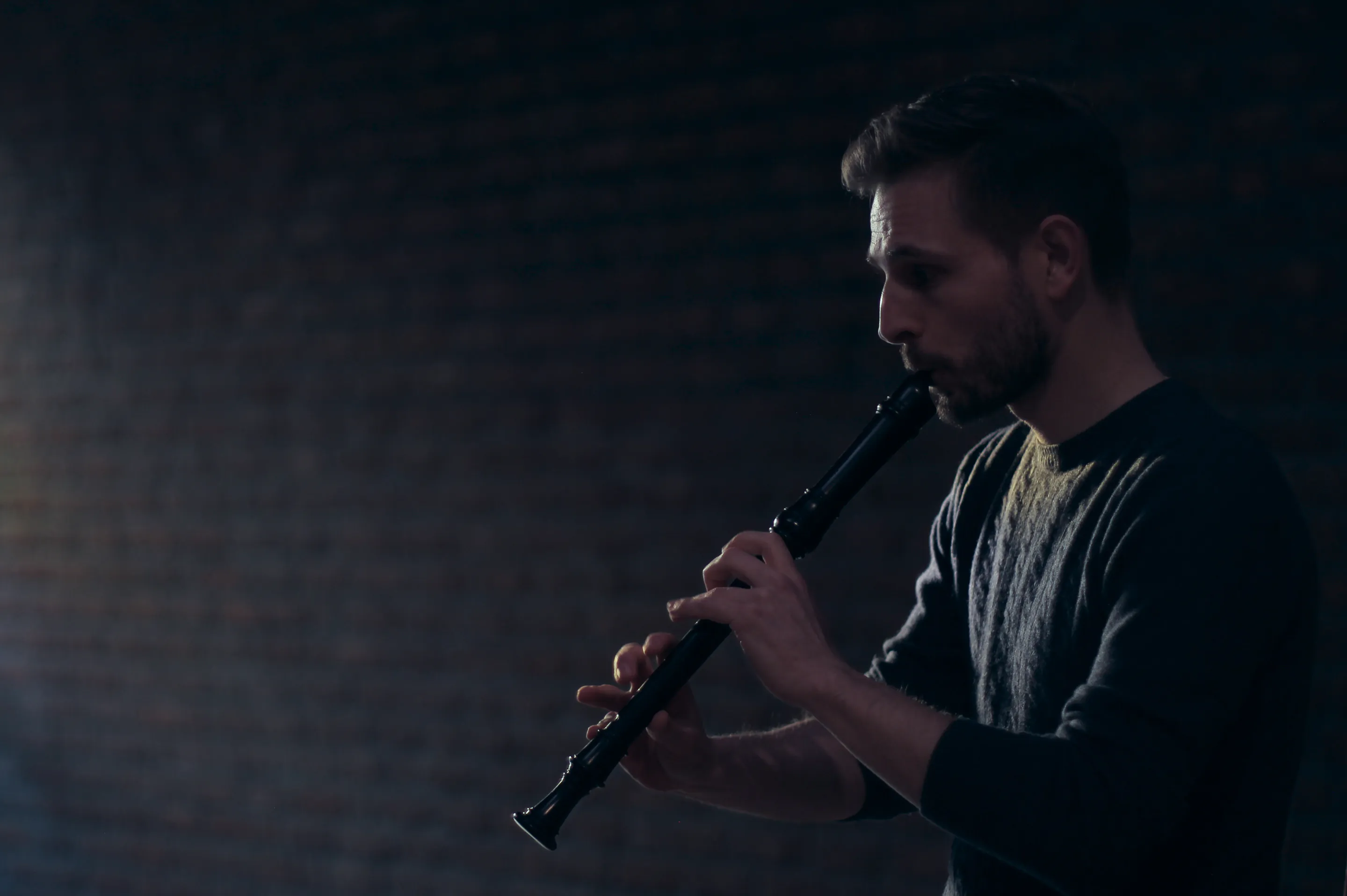 Man playing a black recorder against a dark brick wall background.