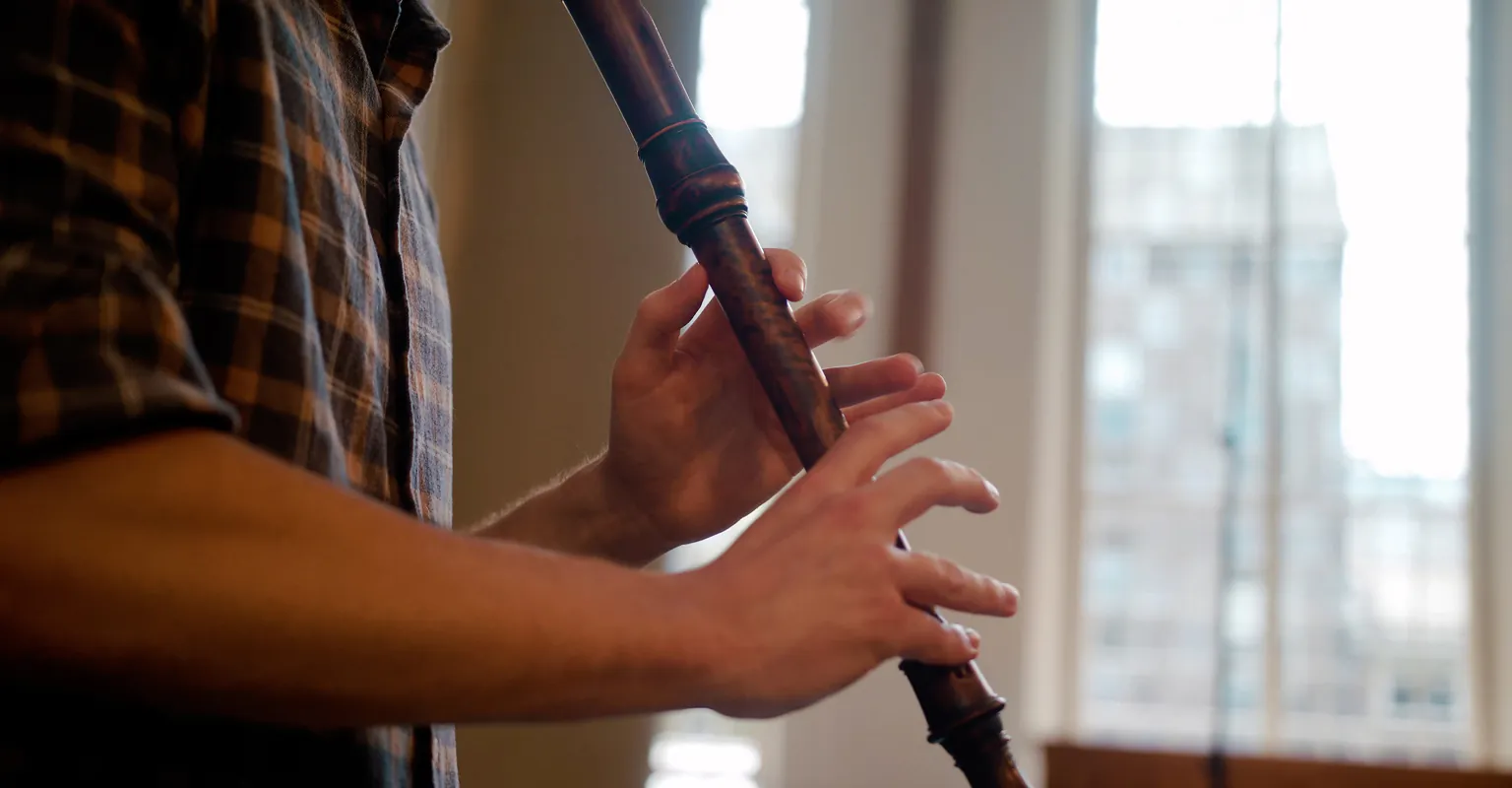 Person playing a recorder indoors near a large window with blurred city view.