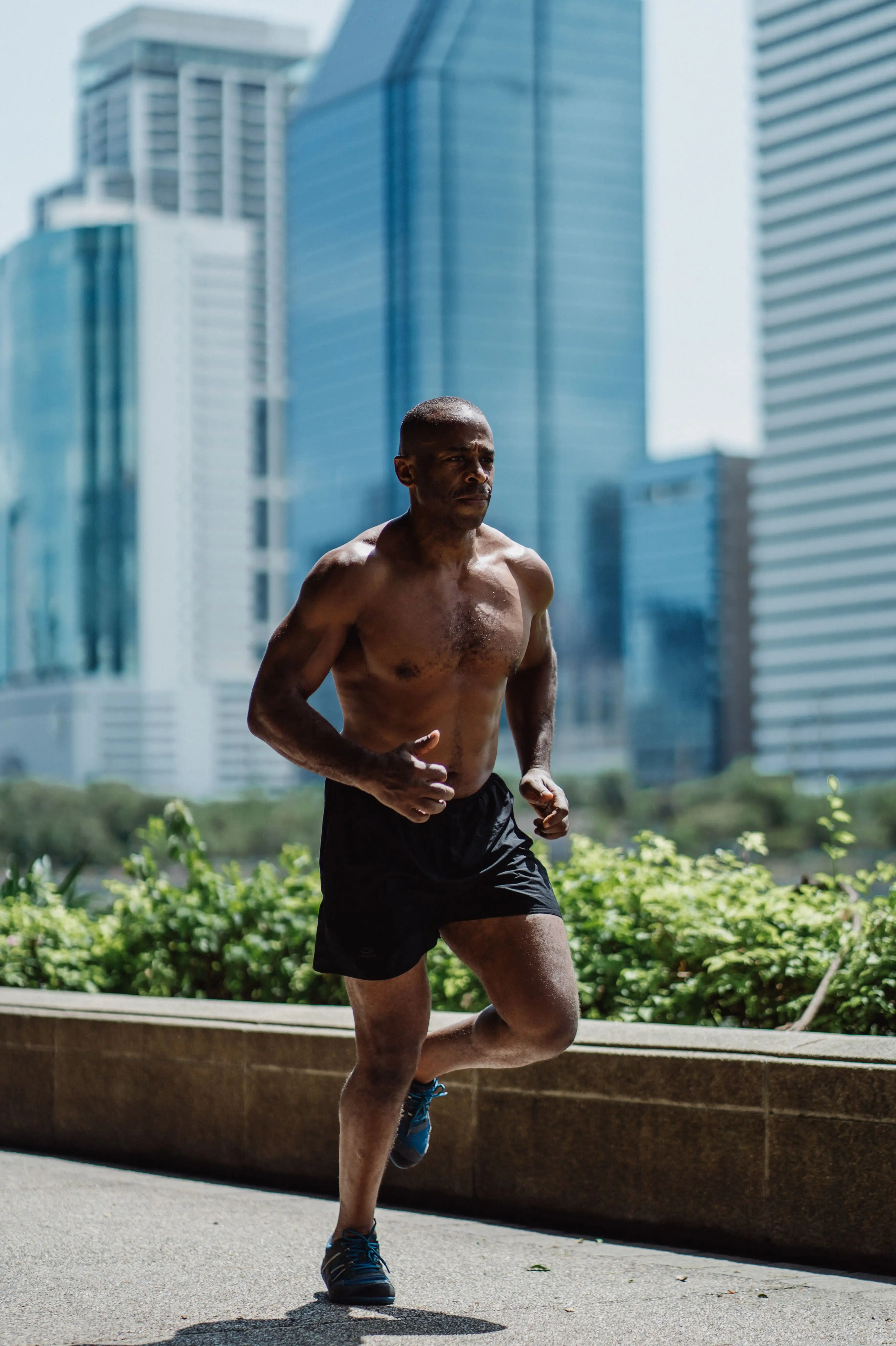 A man with no top on running in the city. there are skyscrapers behind him and he is running on the pavement.