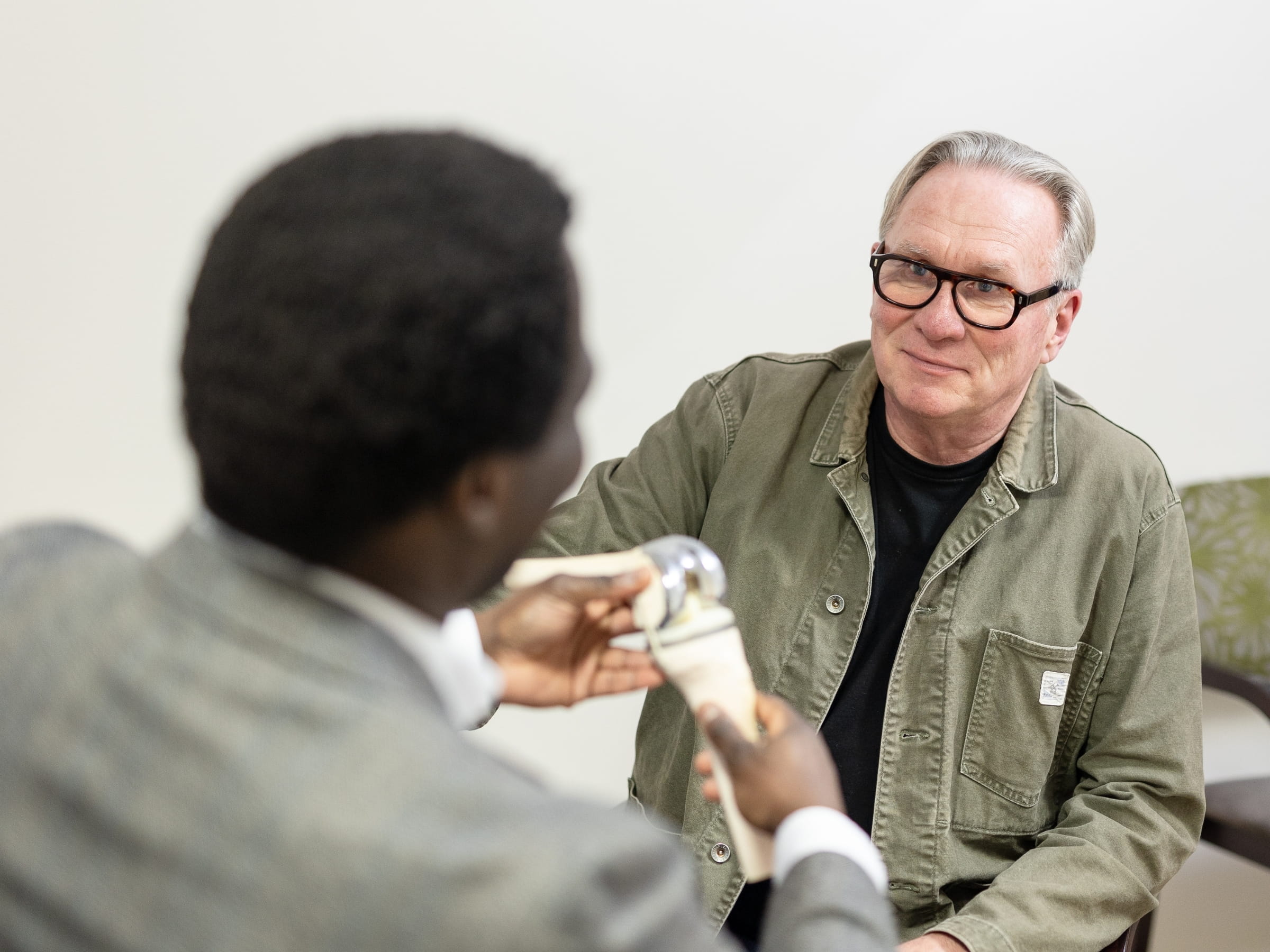 A male patient in his 60s facing the camera in a green jacket, he is talking to mr oni whilst mr oni shows him an articulating model knee joint.
