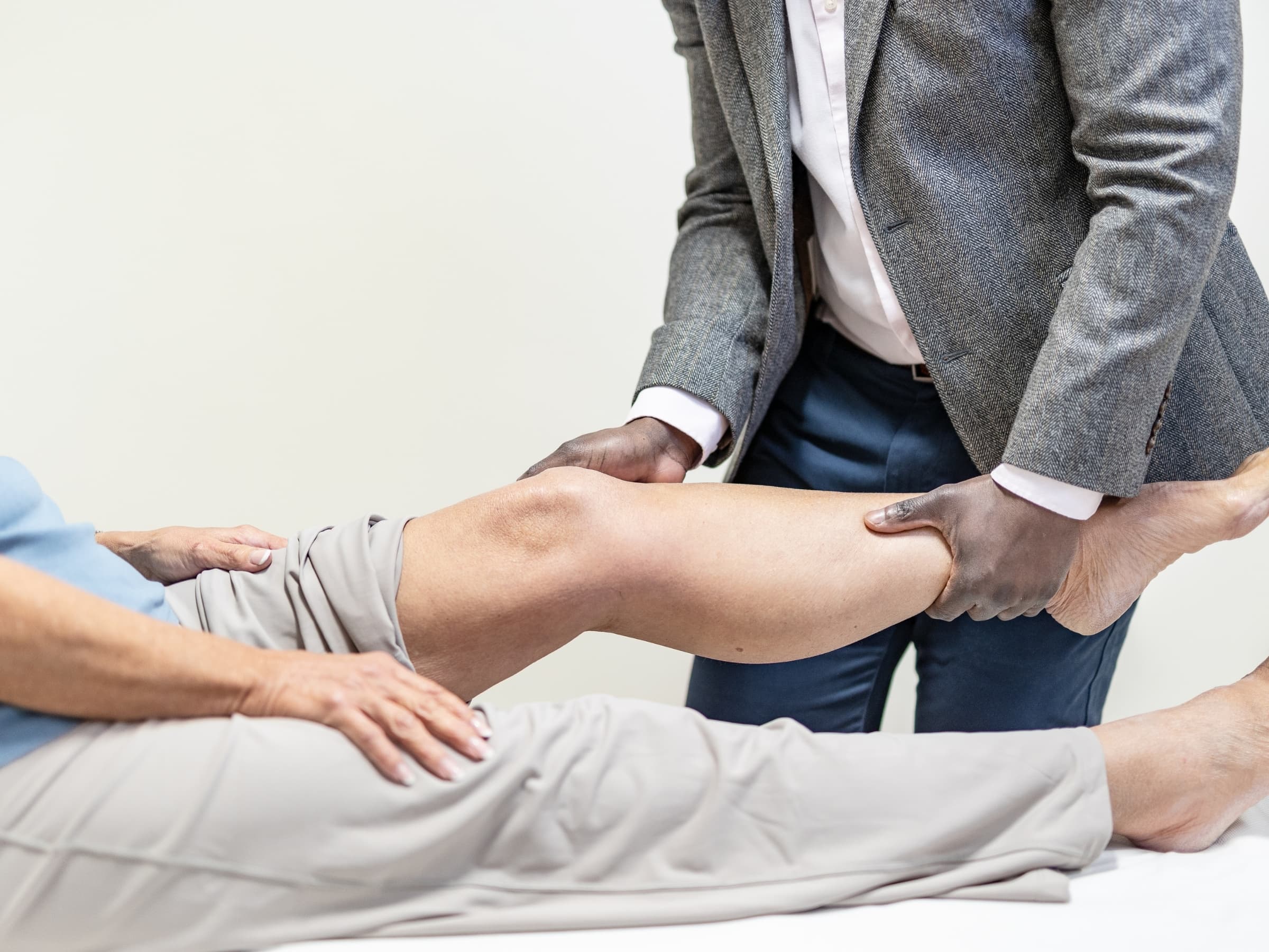 Mr Oni holding the extended leg of a female patient during examination, he is wearing a blazer and standing next to her bed. 
