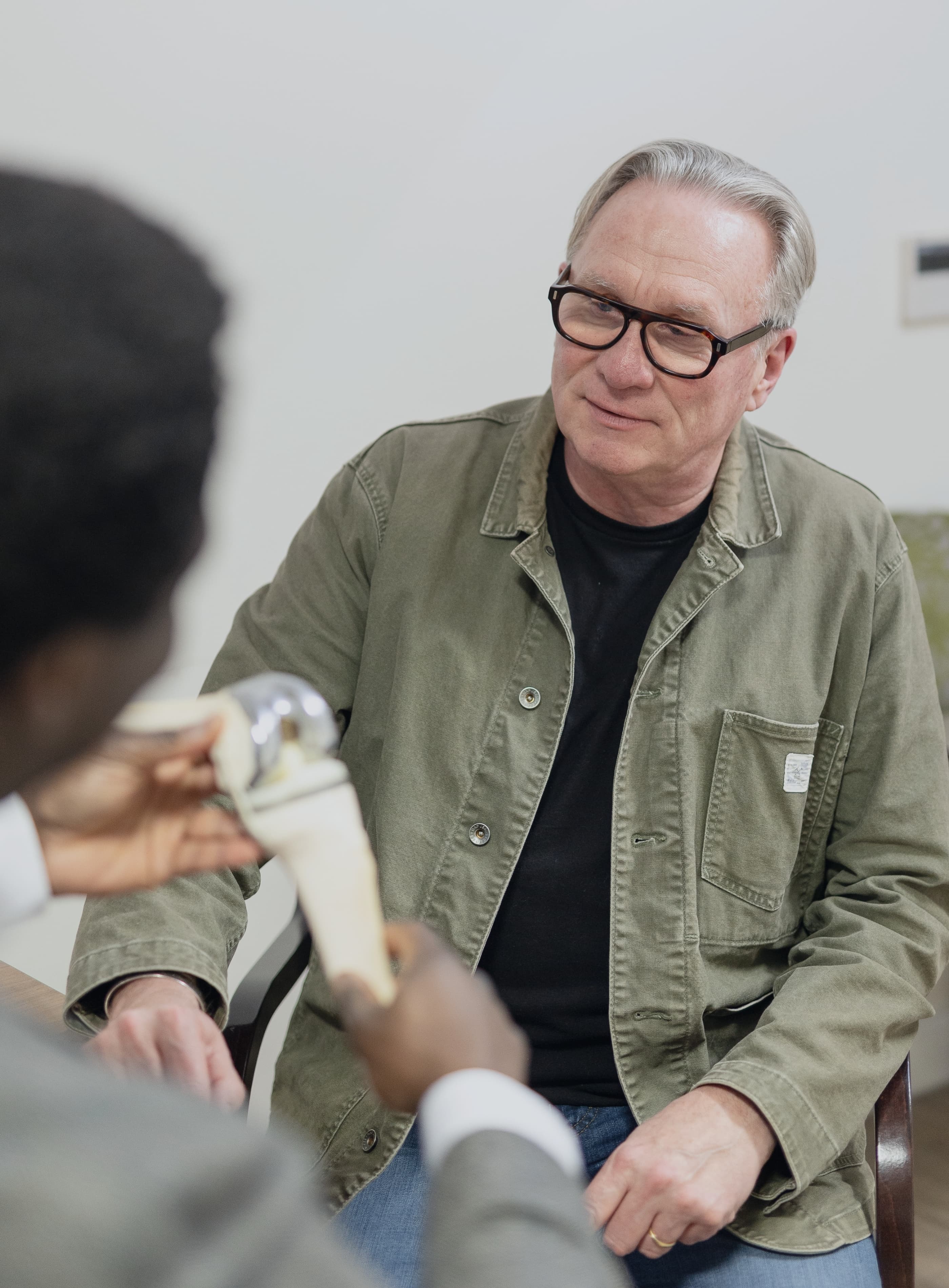 A male patient in a green cargo style jacket looking at Mr Oni as he explains how a knee works with a working model of the knee.