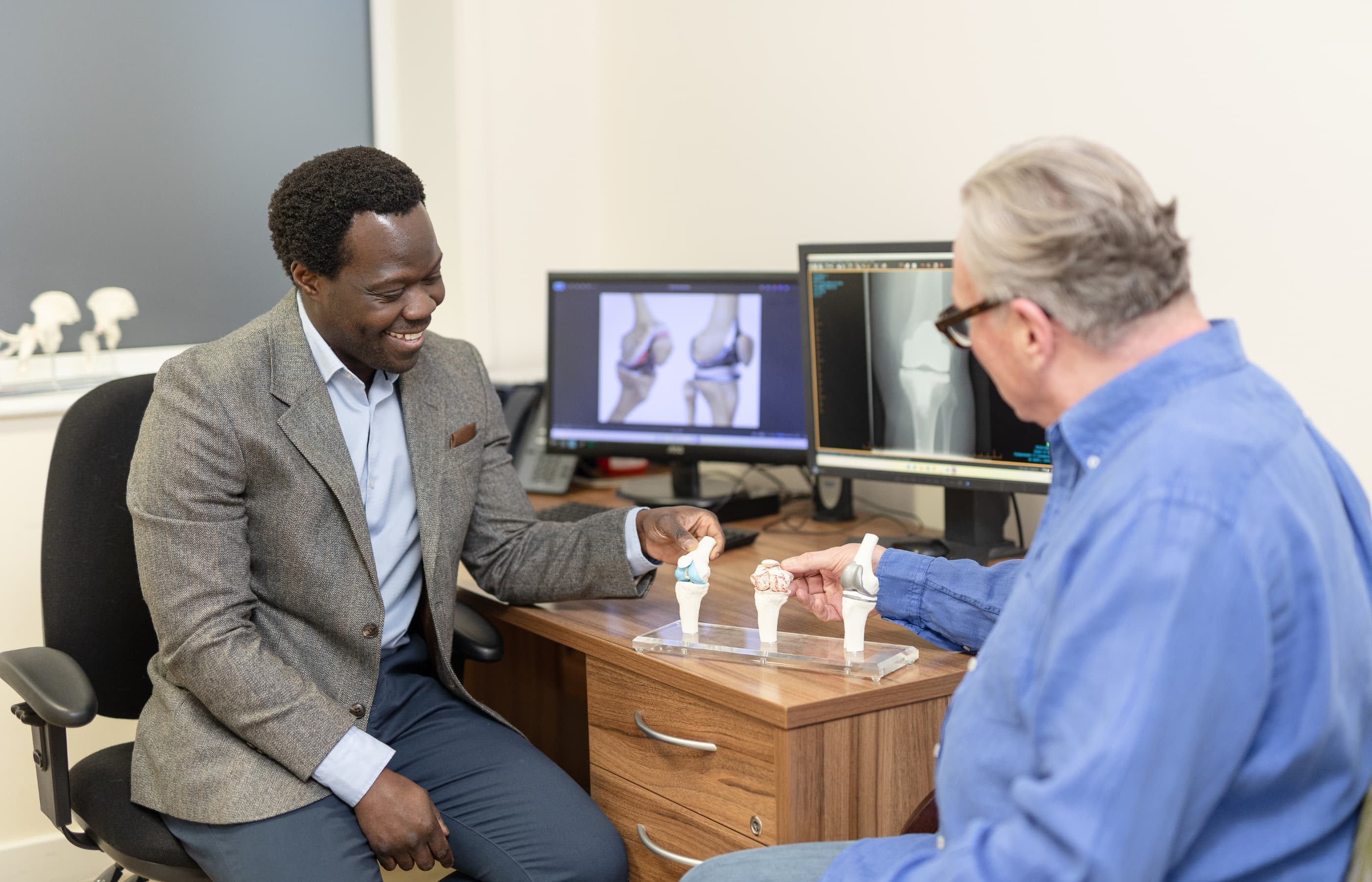 Mr Oni with a male patient who is wearing a blue shirt. they are discussing knee treatment options looking at models of knee joints with varying degrees of knee replacement.