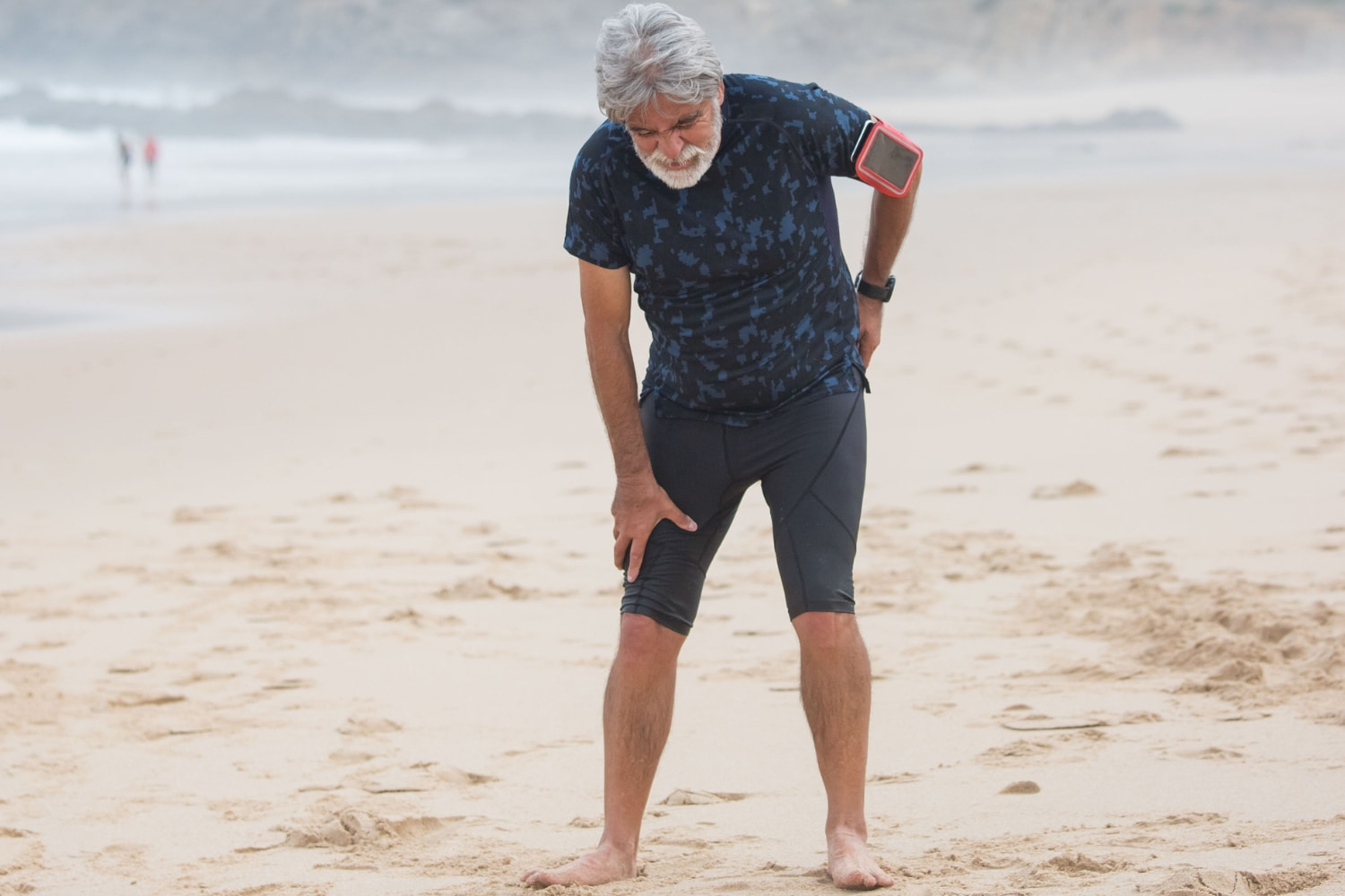 A man on a beach in running gear bending over holding his hip and leg