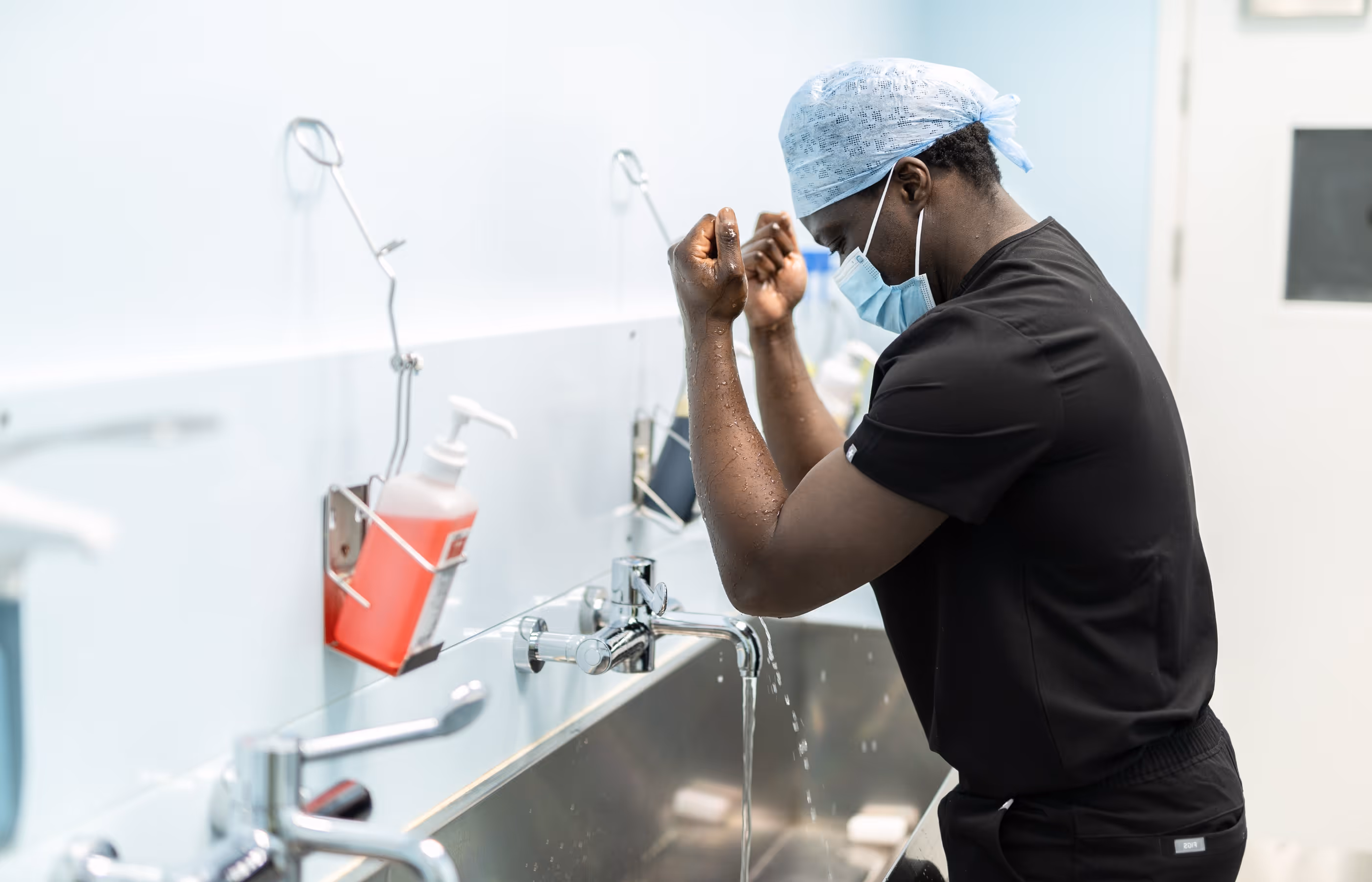 Mr Oni Scrubbing up for surgery in the theatre washing area. He is wearing black scrubs, a mask and a hat.