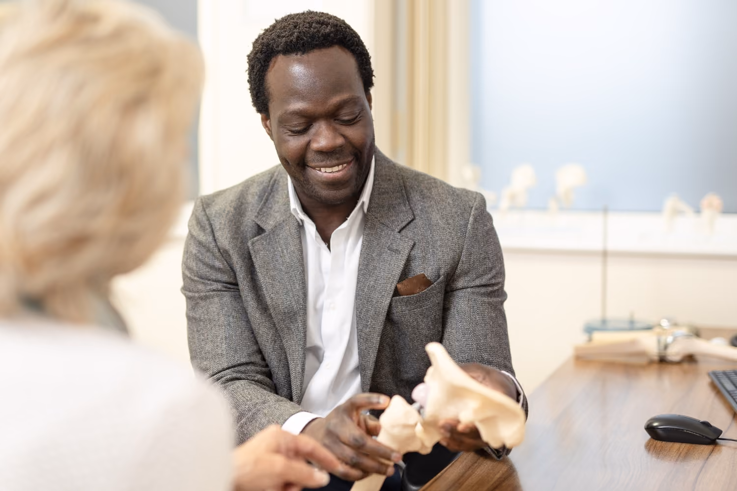 Orthopaedic surgeon in a gray blazer explaining a pelvic bone model to a woman in a medical office.