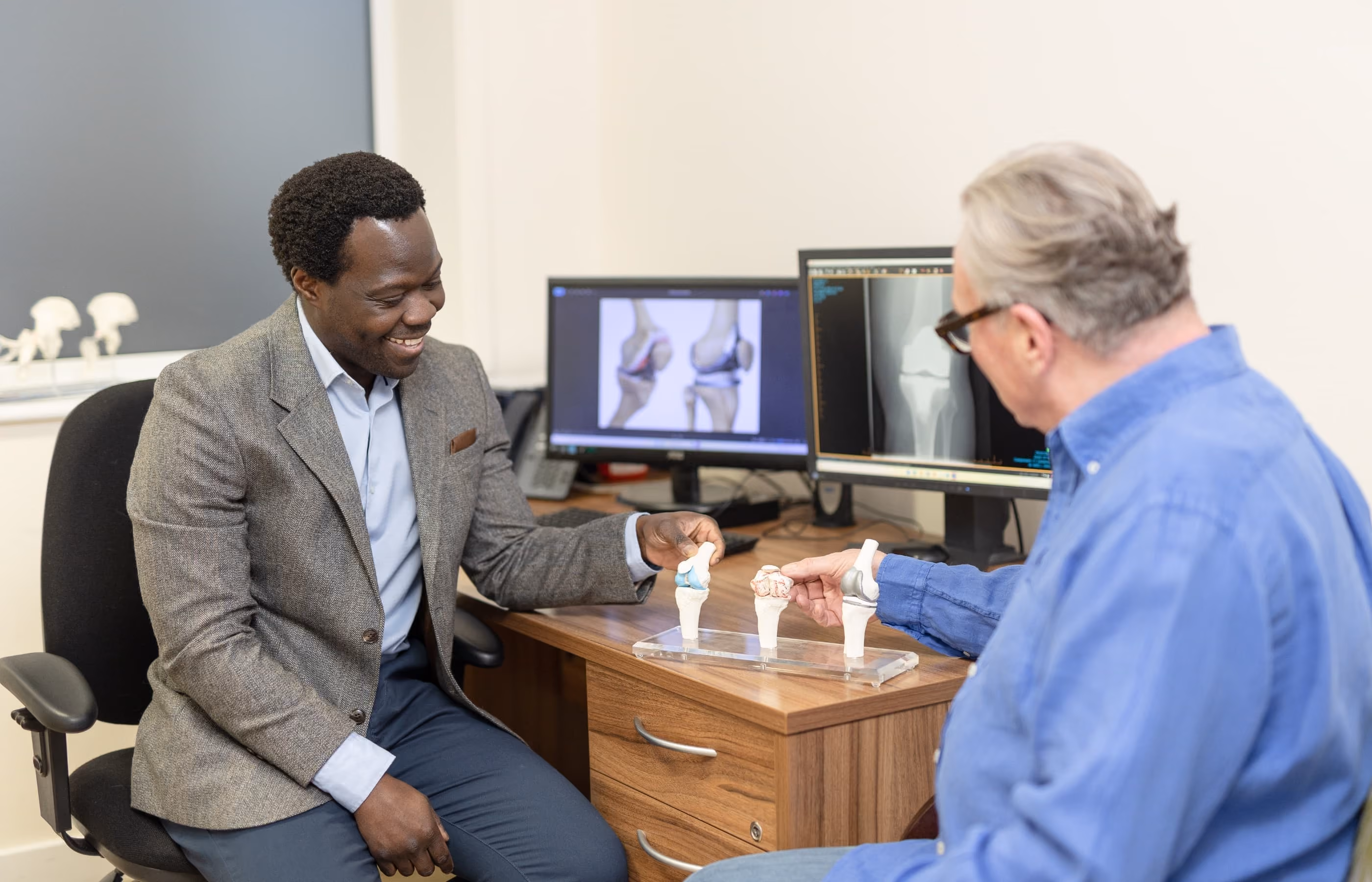 Mr Oni showing a male patient in a blue shirt a medical model of knee joints. They are sat at his desk in front of a computer with x-ray images on the screen.