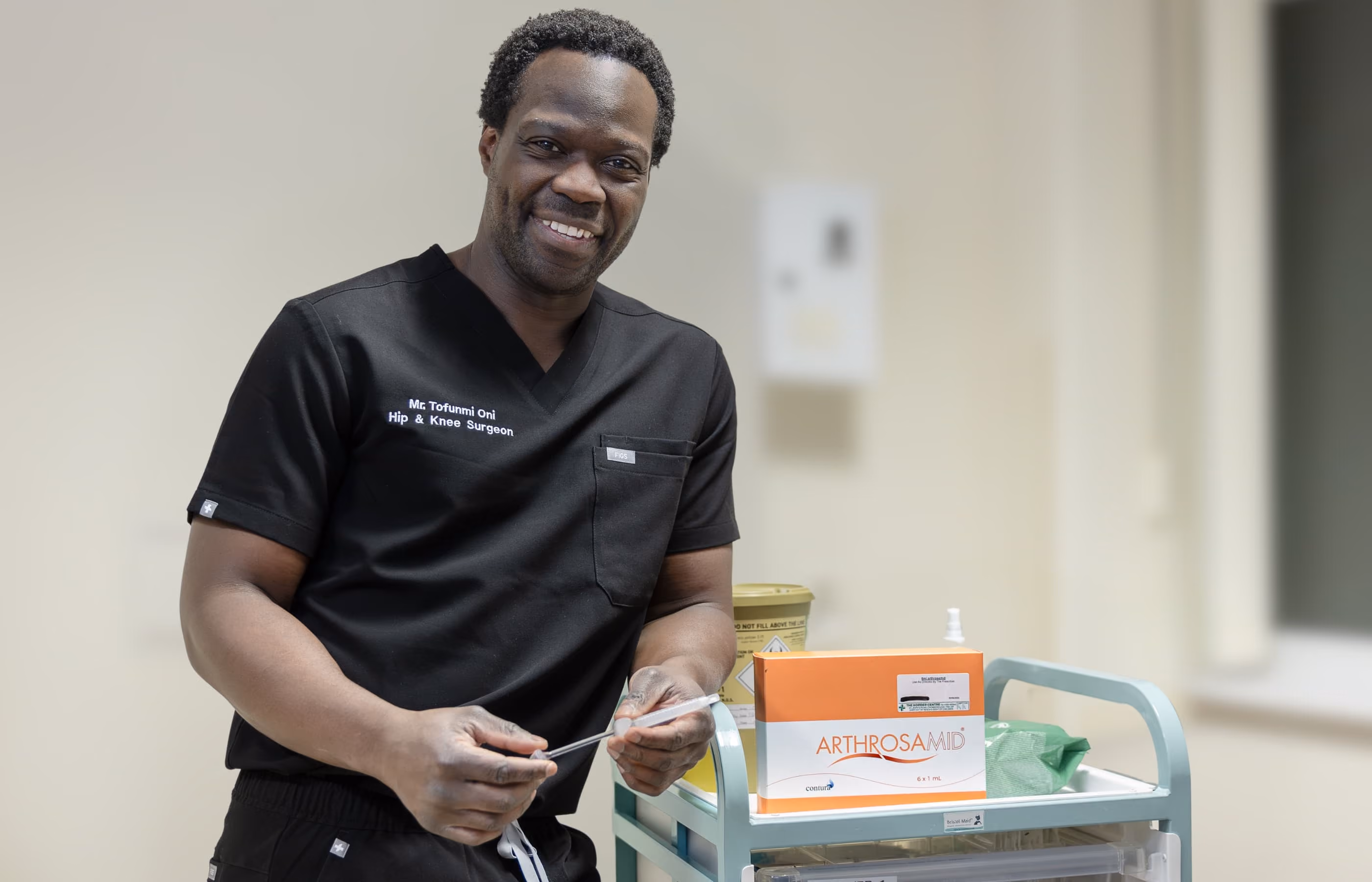 Mr Oni in black scrubs, standing next to a medical trolley with a box of arthrosamid on display