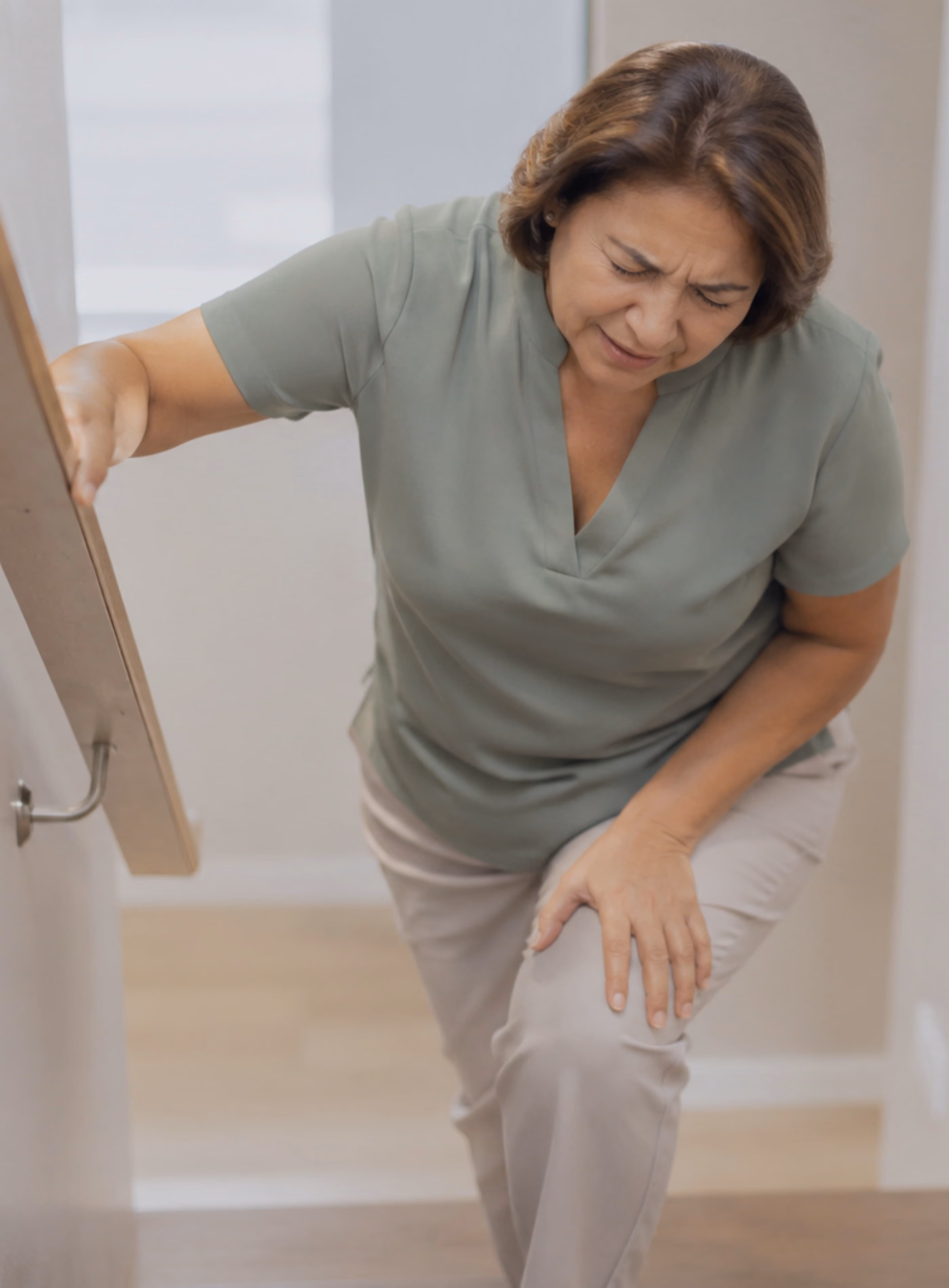 Woman climbing the stairs holding onto the banister, she is grimacing in pain and holding her left knee.
