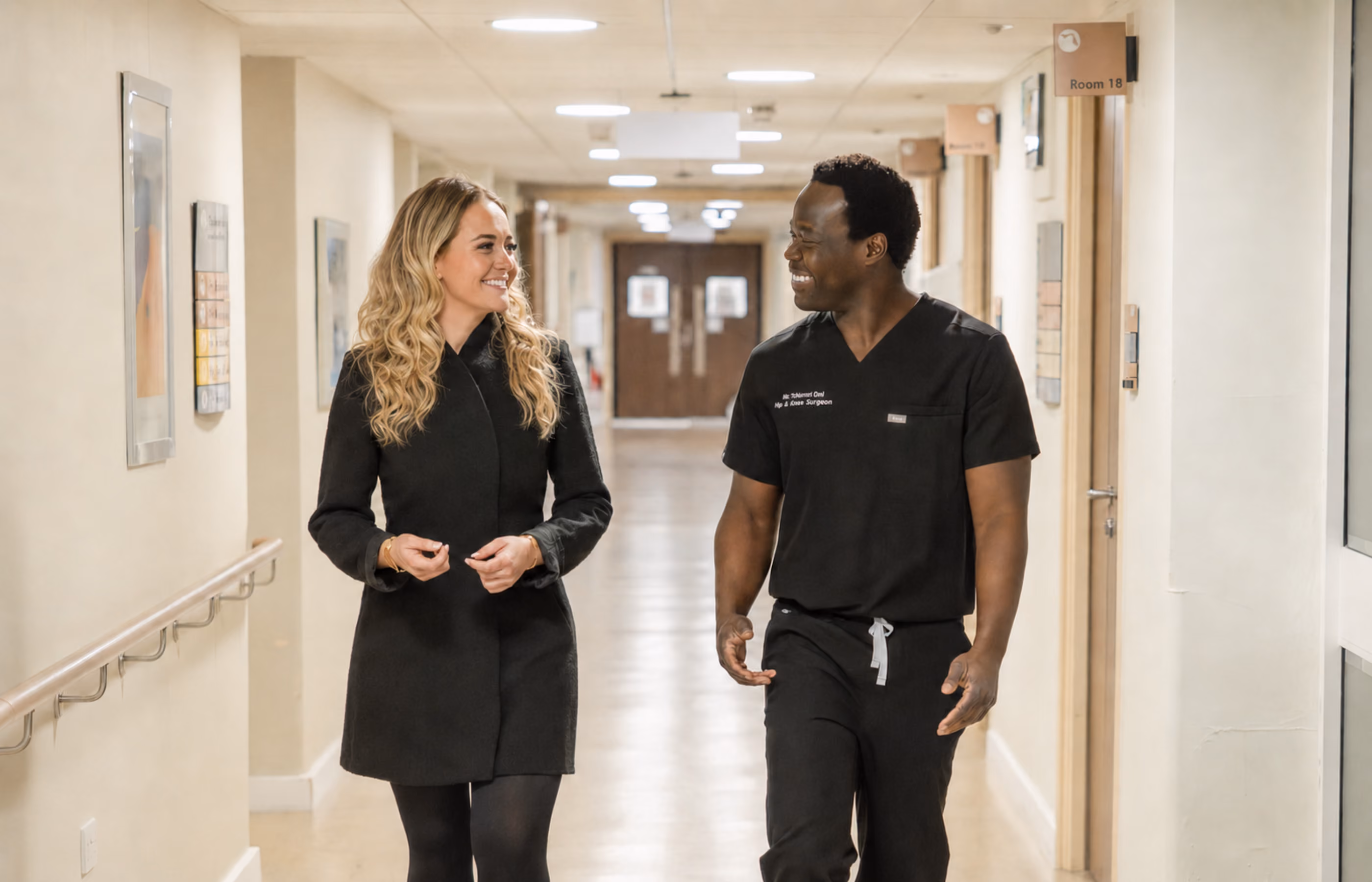 Mr Oni and Libby standing in a hospital corridor talking to each other, smiling and looking please to see each other. 