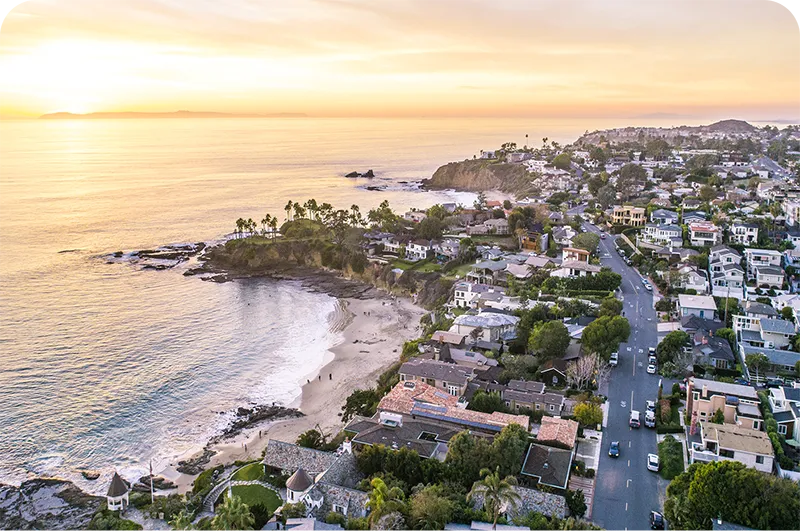 Aerial view of a coastal California town at sunset, with cliffside homes, a sandy beach, and palm trees lining the shore.