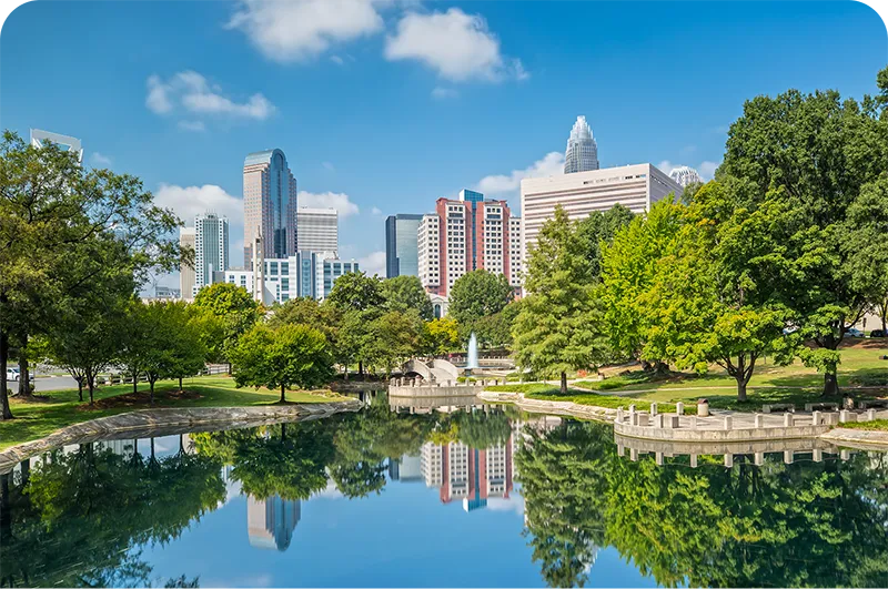 City skyline reflected in a park pond, with trees, a fountain, and a walking path in the foreground.